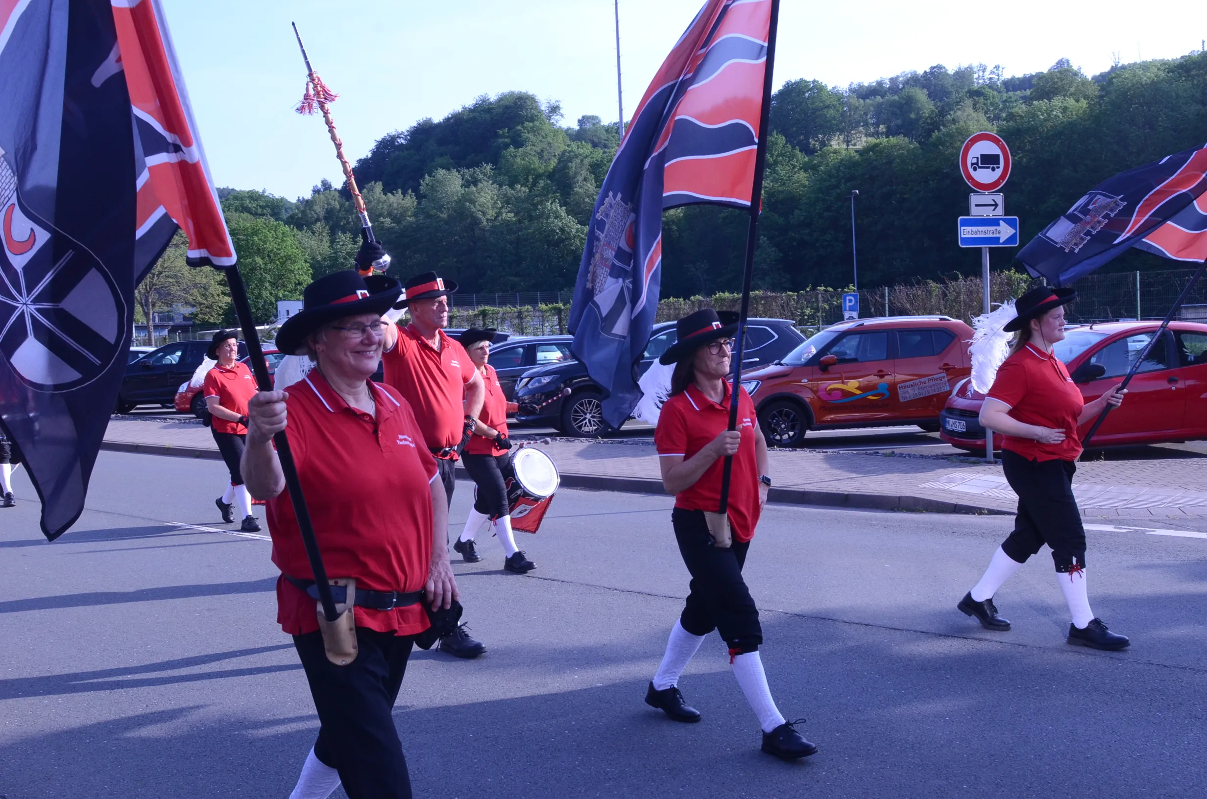 Mehrere Personen in traditioneller Kleidung marschieren mit Fahnen und einer Trommel durch eine Straße, im Hintergrund geparkte Autos und Bäume.