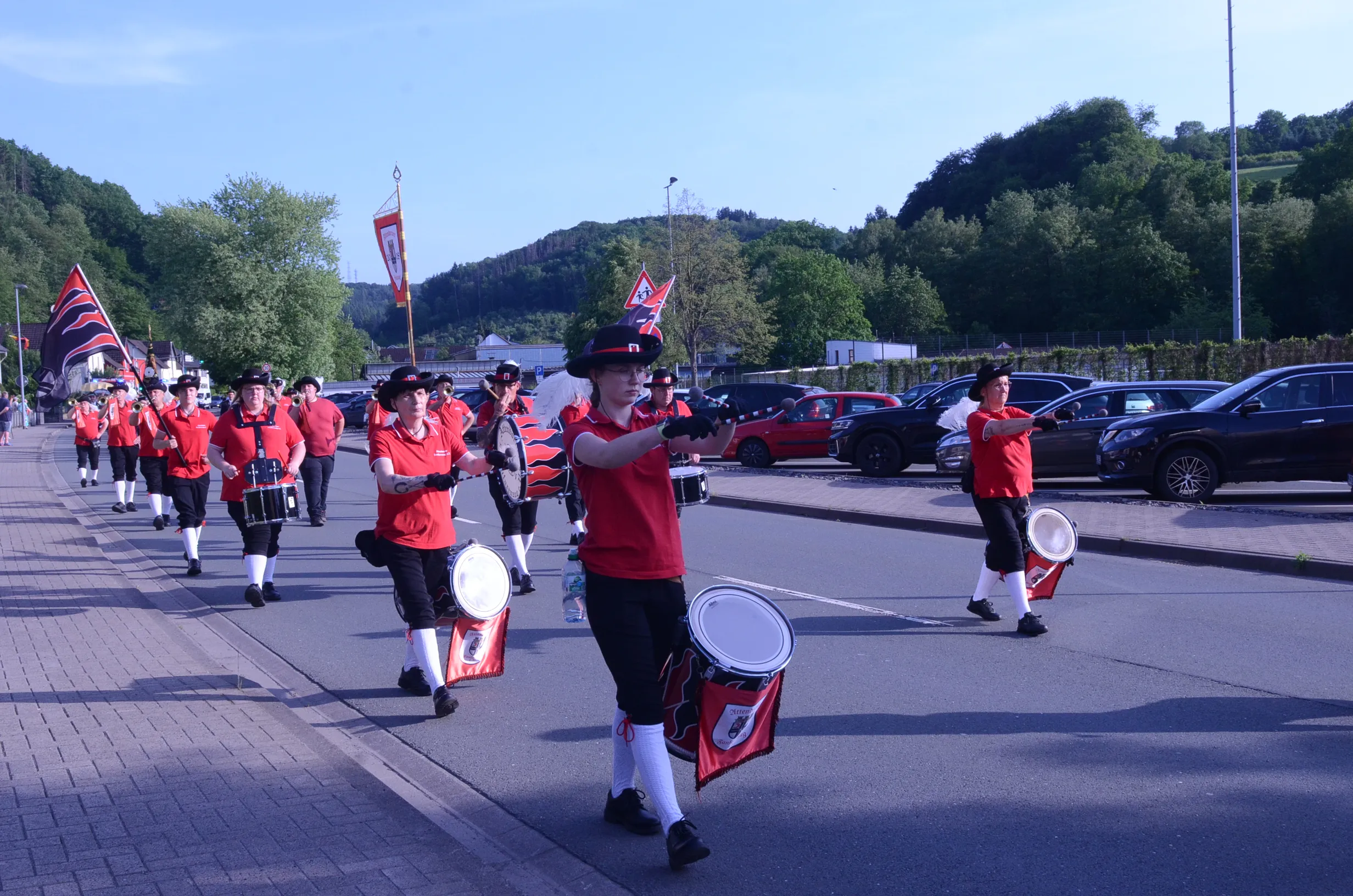 Musikzug in roten Uniformen und mit schwarzen Hüten marschiert auf einer Straße, im Hintergrund parkende Autos und grüne Hügel.