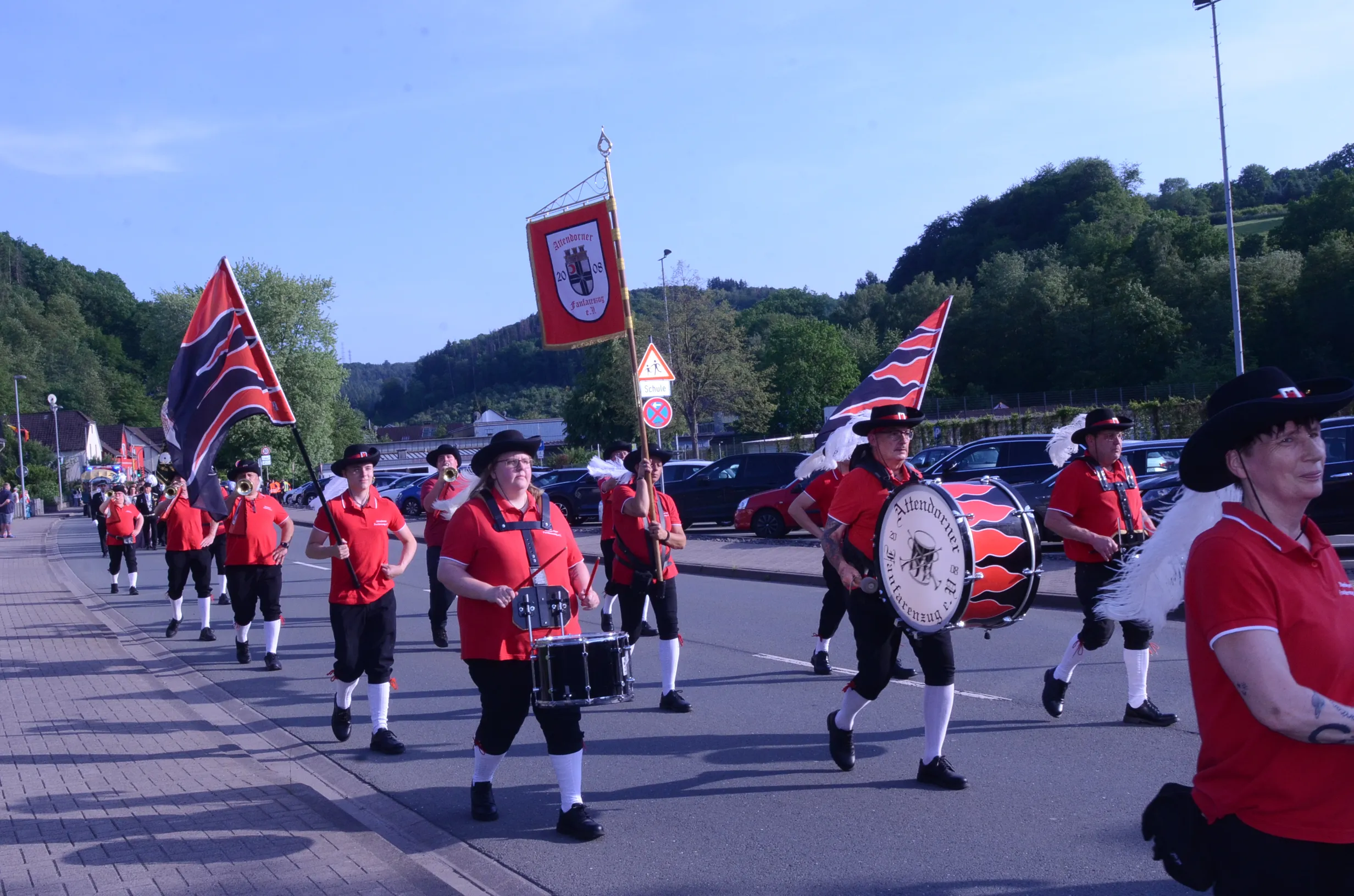 Festzug mit einer Blaskapelle und mehreren Personen in roten Uniformen und schwarzen Hosen. Im Hintergrund eine grüne Hügellandschaft und eine Flagge mit Wappen.