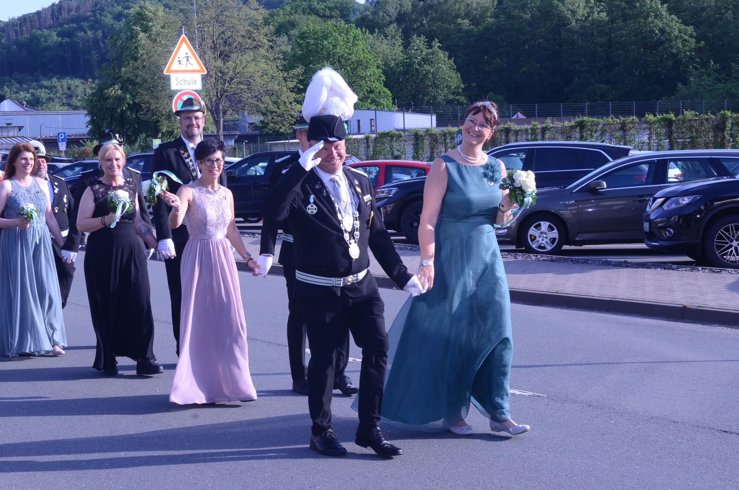 Festzug mit einer festlich gekleideten Gruppe, angeführt von einem Mann in historischer Uniform und einer Frau in Abendkleid, die Hände halten. Im Hintergrund ein Warnschild mit einem Schulkind-Symbol.