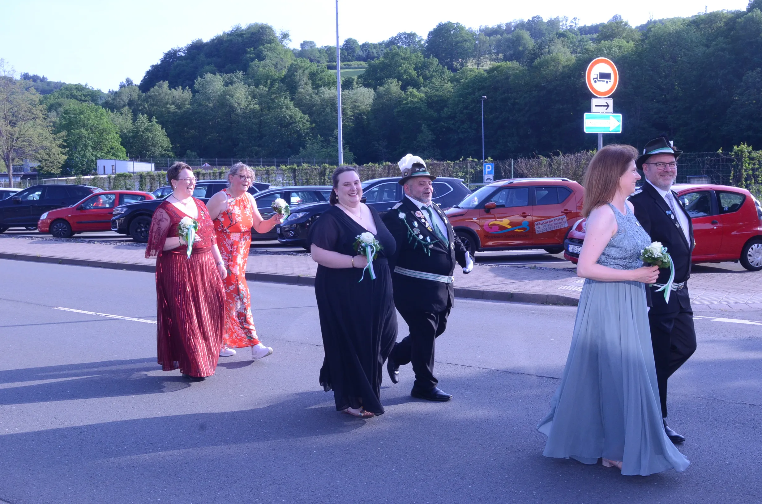 Fünf Personen in festlicher Kleidung gehen auf einem Parkplatz. Die Gruppe besteht aus zwei Frauen in Abendkleidern, einem Mann in Uniform mit Zylinder und zwei weiteren Personen, die ebenfalls festlich gekleidet sind und Blumensträuße tragen.