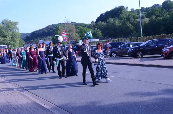Festzug mit Schützen in historischen Uniformen und festlich gekleideten Begleitern, darunter eine Frau, die winkt. Im Hintergrund geparkte Autos und eine bewaldete Hügelkette.