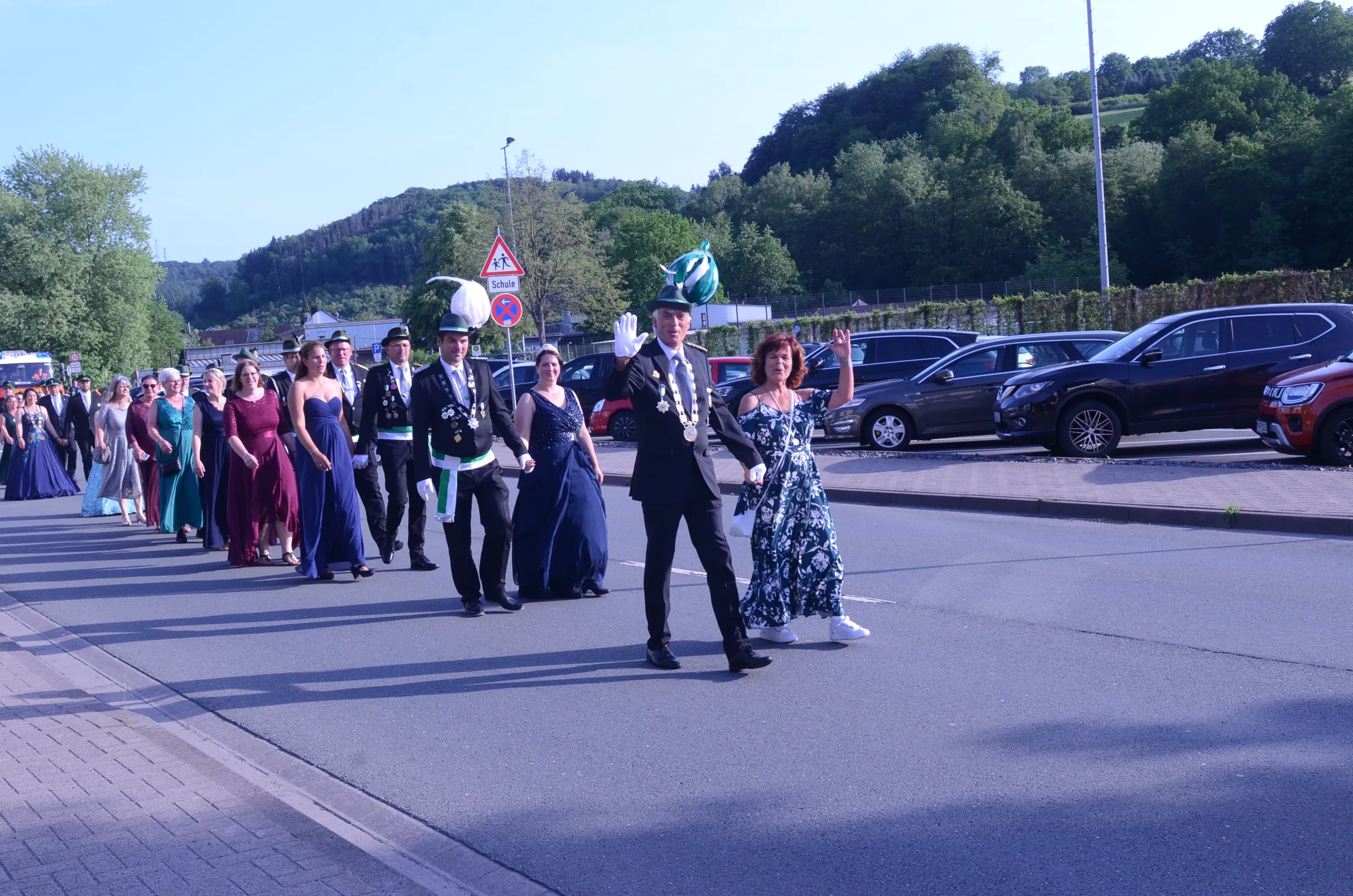 Festzug mit Schützen in historischen Uniformen und festlich gekleideten Begleitern, darunter eine Frau, die winkt. Im Hintergrund geparkte Autos und eine bewaldete Hügelkette.