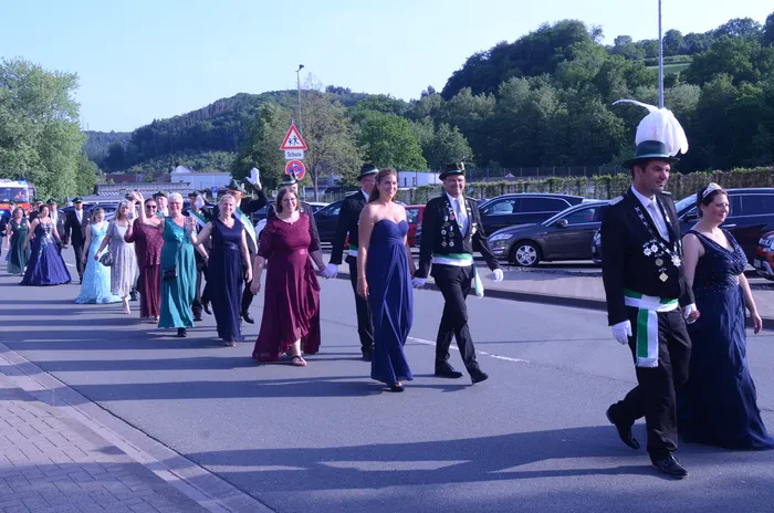 Festzug mit festlich gekleideten Paaren und einer Blaskapelle, die eine Straße entlanggehen. Im Hintergrund sind Zuschauer und geparkte Autos zu sehen.