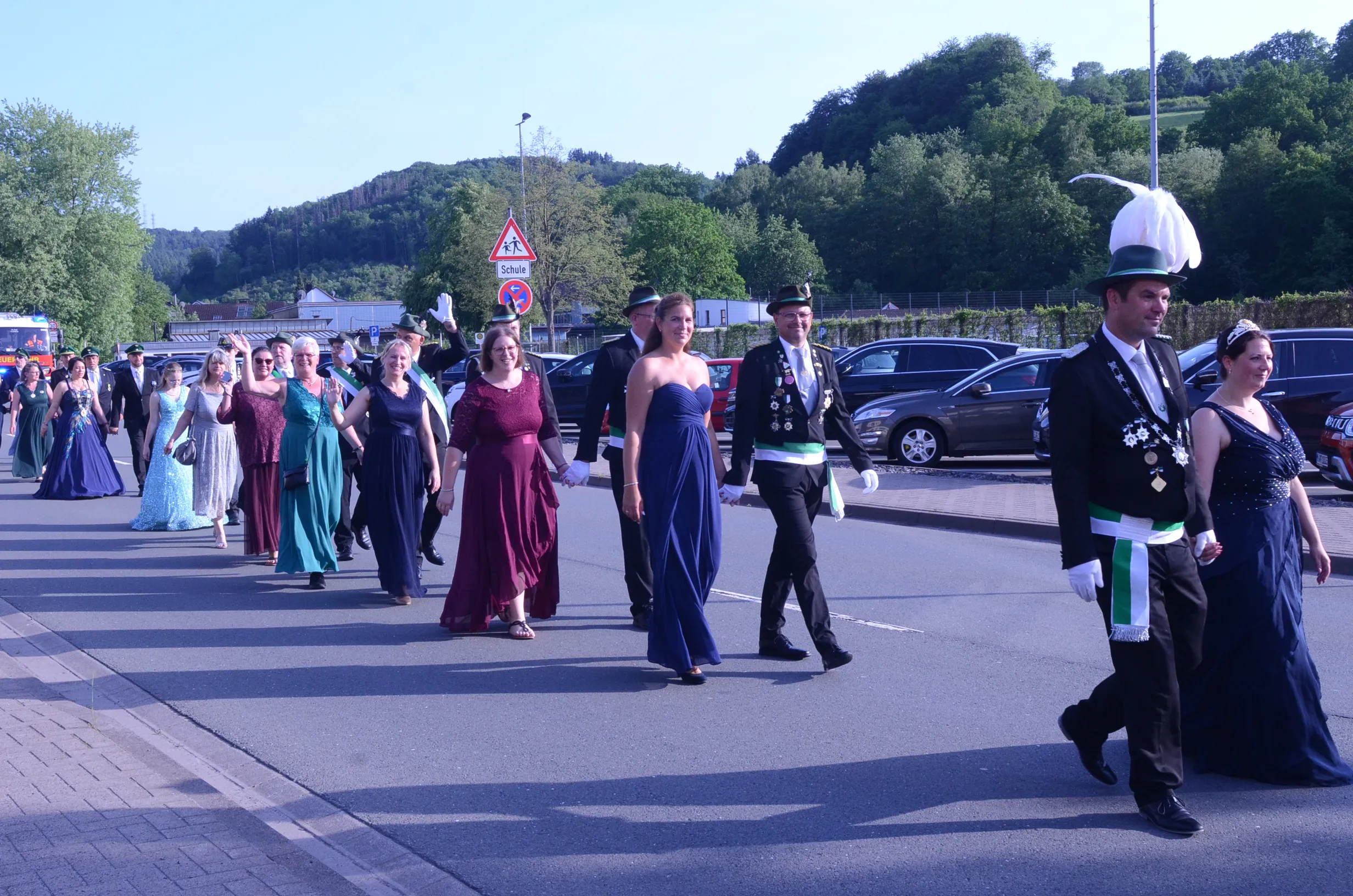 Festzug mit festlich gekleideten Paaren und einer Blaskapelle, die eine Straße entlanggehen. Im Hintergrund sind Zuschauer und geparkte Autos zu sehen.