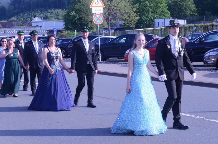 Ein Jugendpaar in Festkleidung schreitet Hand in Hand vor einer Gruppe von Begleitern in Uniformen und Abendgarderobe durch eine Straße. Im Hintergrund ein Straßenschild mit dem Hinweis auf eine Schule.