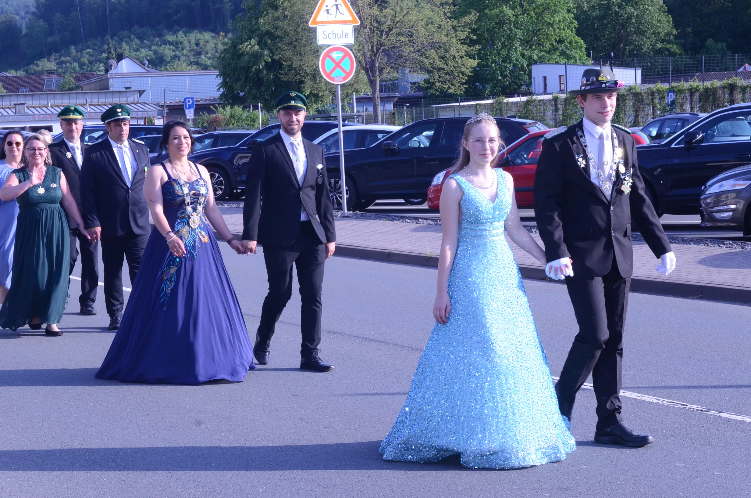 Ein Jugendpaar in Festkleidung schreitet Hand in Hand vor einer Gruppe von Begleitern in Uniformen und Abendgarderobe durch eine Straße. Im Hintergrund ein Straßenschild mit dem Hinweis auf eine Schule.