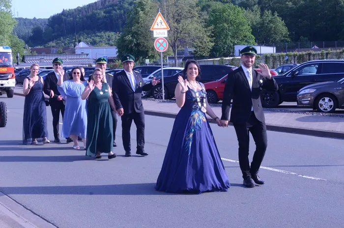 Paar in Festkleidung hält Hände und winkt während eines Umzugs, begleitet von weiteren Personen in Uniformen und langen Kleidern. Im Hintergrund ein Straßenschild mit einem Schulkind-Symbol.