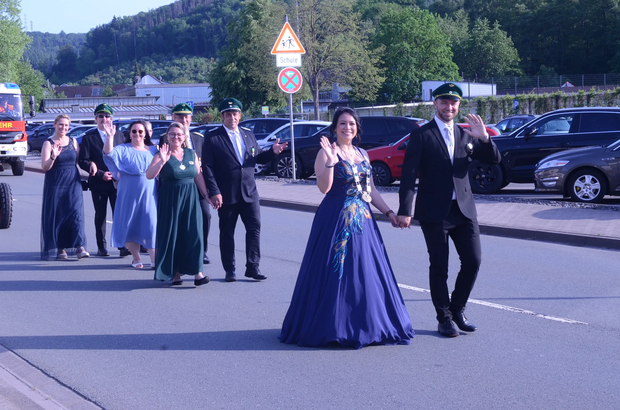 Paar in Festkleidung hält Hände und winkt während eines Umzugs, begleitet von weiteren Personen in Uniformen und langen Kleidern. Im Hintergrund ein Straßenschild mit einem Schulkind-Symbol.