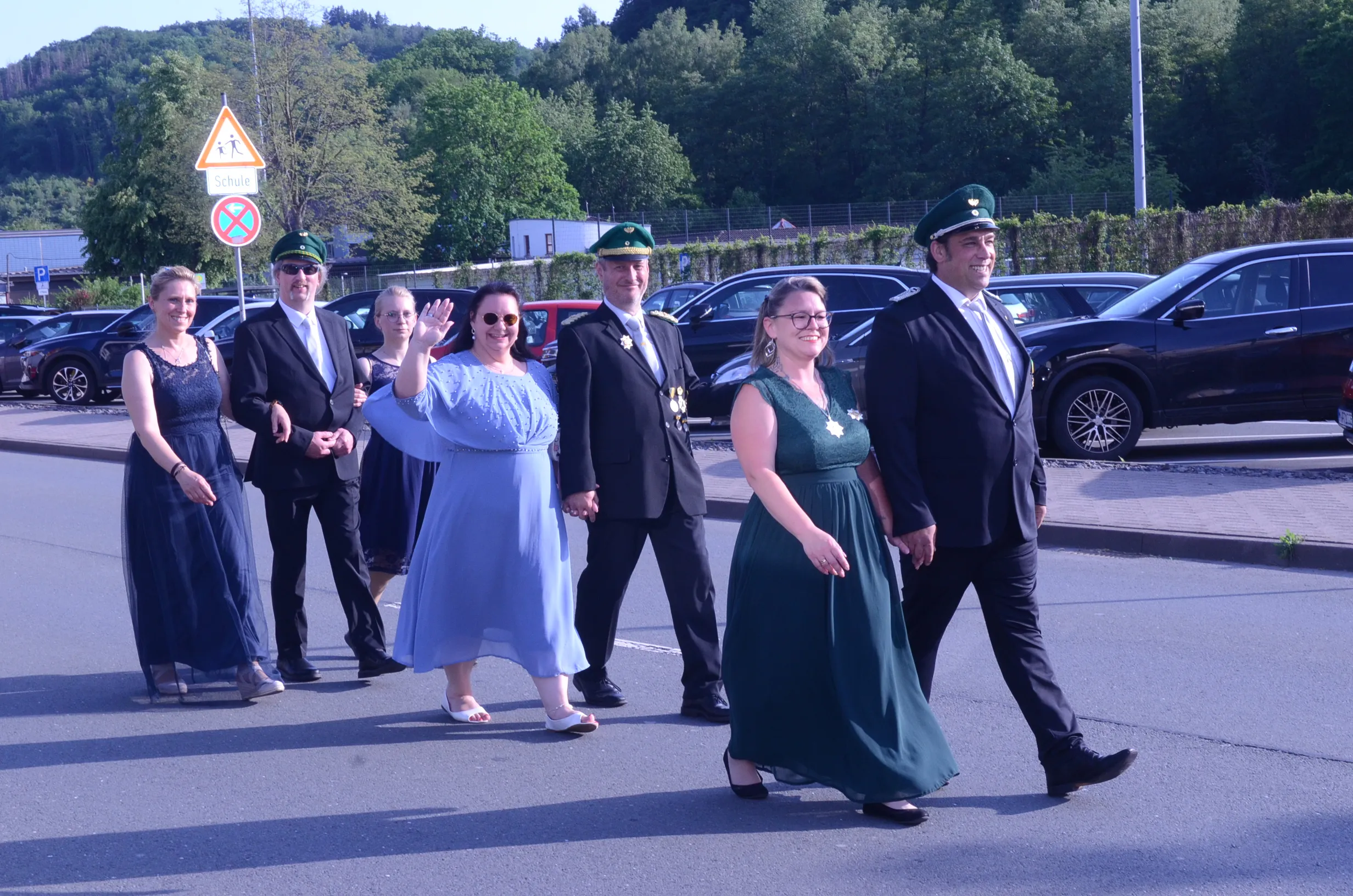 Festzug mit mehreren Personen in festlicher Kleidung, darunter uniformierte Mitglieder einer Schützenkompanie, schreitet über eine Straße. Im Hintergrund ein Verkehrsschild mit dem Hinweis auf eine Schule.