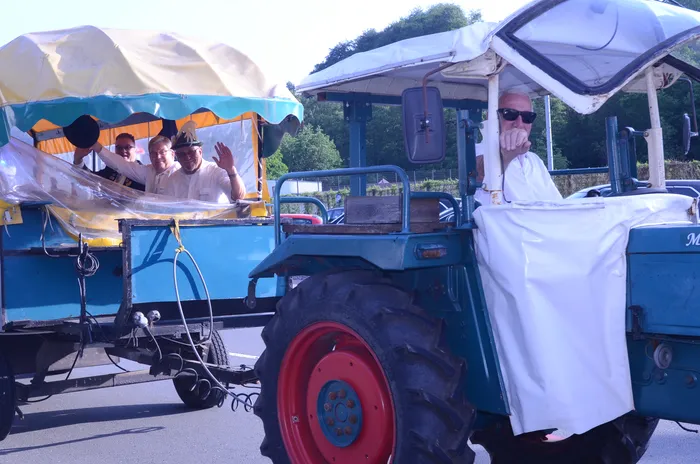 Zwei restaurierte Oldtimer-Traktoren fahren in einem Festumzug, jeweils mit mehreren Personen, die zum Teil winken. Die Traktoren sind blau und mit weißen Planen verziert.