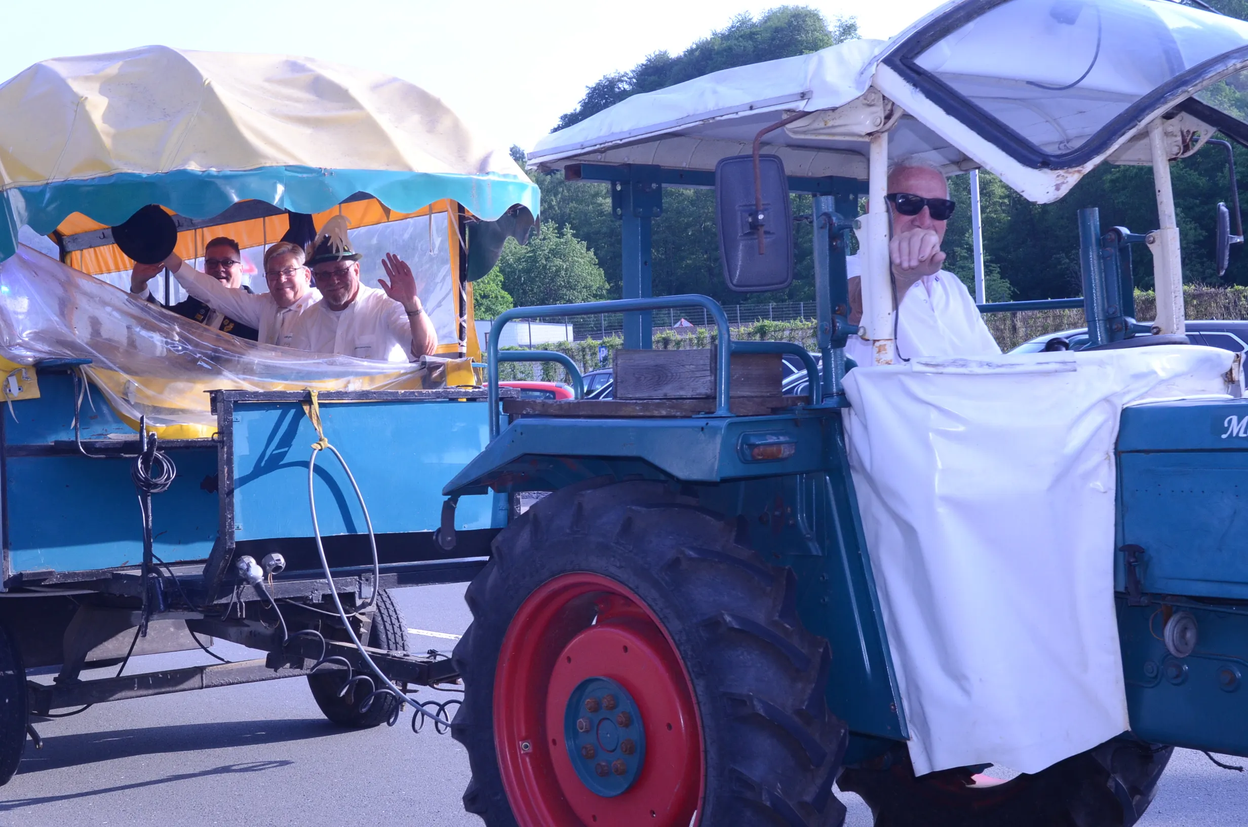 Zwei restaurierte Oldtimer-Traktoren fahren in einem Festumzug, jeweils mit mehreren Personen, die zum Teil winken. Die Traktoren sind blau und mit weißen Planen verziert.