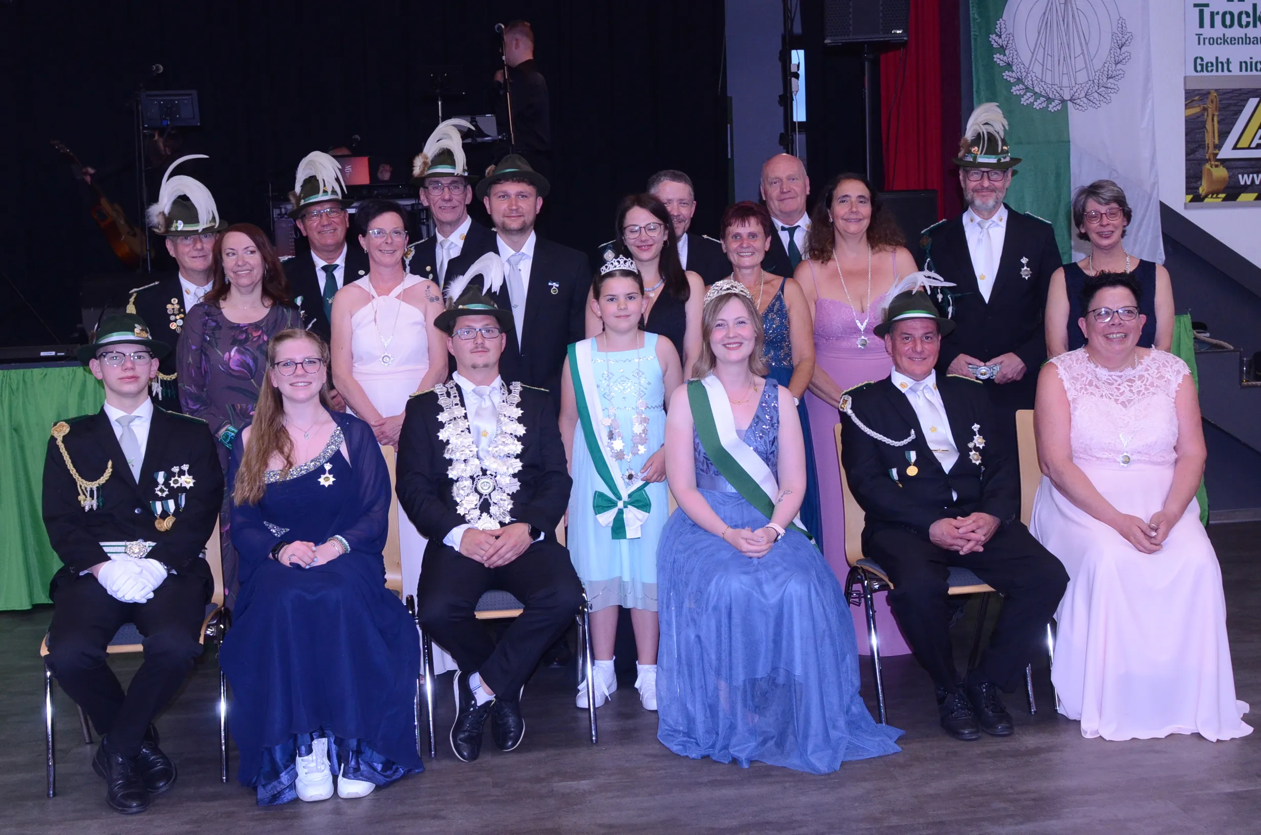 Gruppenfoto von etwa zwanzig Personen in festlicher Kleidung, darunter traditionelle Schützenuniformen und Abendkleider, vor einem Bühnenhintergrund.