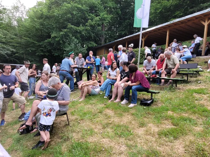 Zuschauer sitzen und stehen auf einer Rasenfläche vor Holzbänken und Tischen, während ein Mann mit einem Gewehr im Vordergrund zielt. Im Hintergrund sind Bäume und eine grüne Flagge zu sehen.