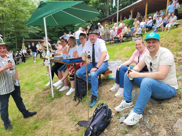Menschen sitzen und stehen auf einer Rasenfläche vor einer Holzhütte, einige halten Getränke in der Hand. Im Hintergrund ein grüner Schirm und weitere Zuschauer.