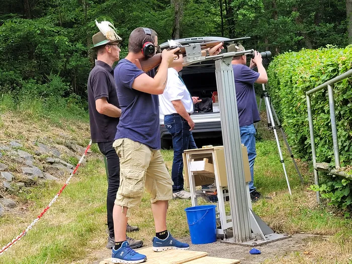 Ein Schütze zielt mit einem Gewehr auf eine Zielscheibe, die an einer Metallkonstruktion befestigt ist. Im Hintergrund stehen weitere Personen und ein Lieferwagen.