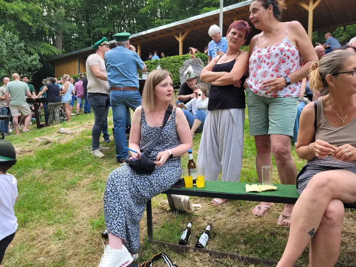 Menschen stehen und sitzen auf einer Wiese vor einem Holzpavillon, einige halten Bierflaschen. Im Hintergrund sind weitere Personen und Bäume zu sehen.