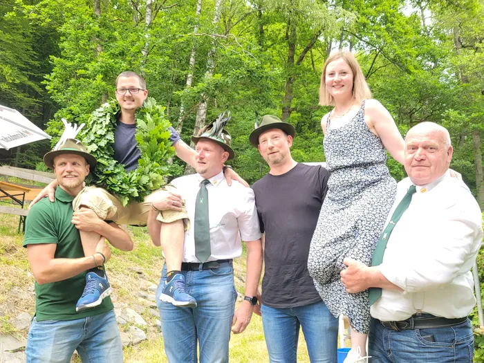 Fünf lächelnde Personen stehen im Freien vor einer grünen Hecke. Drei Männer tragen traditionelle Schützenkleidung mit grüfen Hüten und orange-goldenen Bändern, zwei weitere Personen tragen Freizeitkleidung. Einer der Männer hält einen großen Zweig mit Blättern.