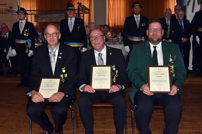 Drei Männer in dunkler Kleidung sitzen auf einer Bühne und halten Urkunden. Sie tragen Schützenorden und Medaillen. Im Hintergrund stehen weitere Personen in Schützenuniformen.