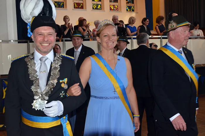Ein Mann und eine Frau lächeln in die Kamera. Der Mann trägt eine dunkle Uniform mit vielen Orden und eine Kette, die Frau ein hellblaues Kleid und eine Tiara. Im Hintergrund sind weitere Personen und eine Fensterfront mit bunten Glasfenstern zu sehen.