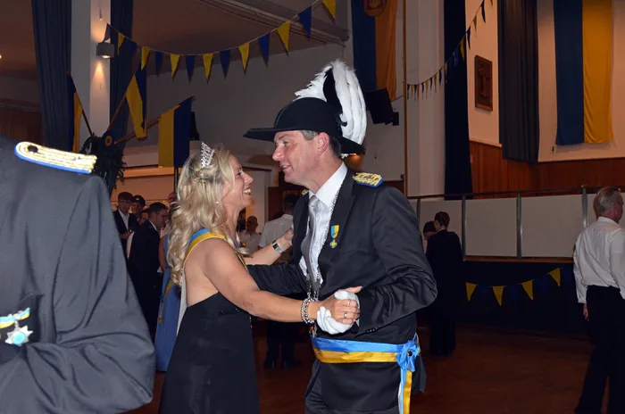 Ein Mann in Uniform und Dreispitz tanzt mit einer Frau in Abendkleid und Tiara in einem festlich dekorierten Saal. Im Hintergrund sind weitere tanzende Personen und Girlanden in Gelb und Blau zu sehen.