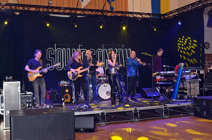 Eine Band mit sechs Mitgliedern spielt auf einer Bühne vor einem dunklen Hintergrund mit dem Schriftzug „Soundconvoy“. Die Musiker spielen verschiedene Instrumente wie Gitarren, Bass, Schlagzeug und Keyboard.