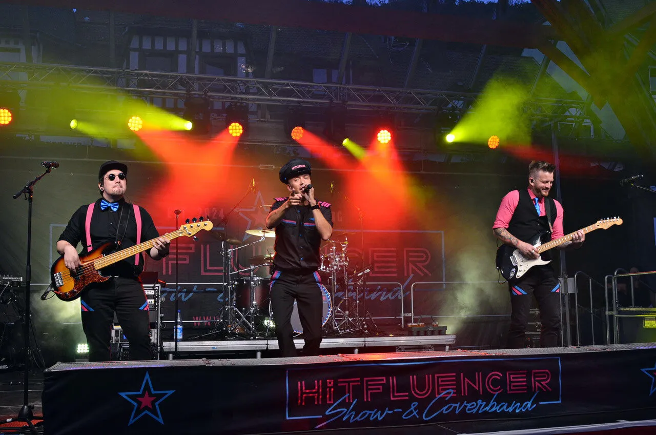 Die Band Hitfluencer tritt live auf einer Bühne unter gelben und roten Scheinwerfern auf. Zu sehen sind ein Bassist, ein Sänger und ein Gitarrist.