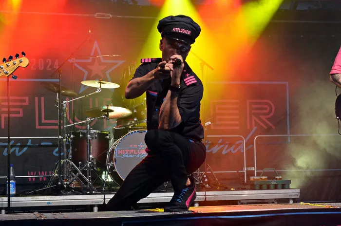Ein Sänger der Band Hitfluencer in schwarzer Uniform mit pinken Akzenten singt auf einer beleuchteten Bühne in ein Mikrofon.
