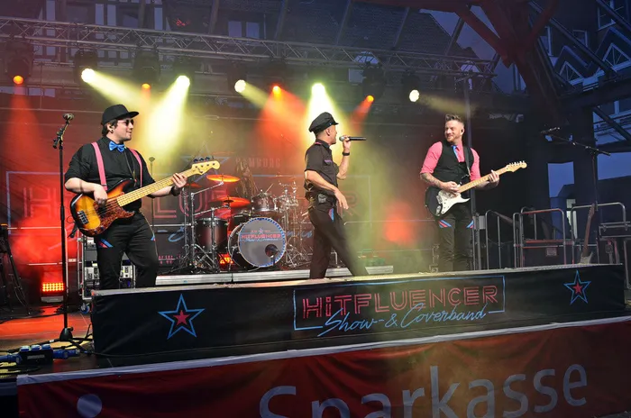 Die Band Hitfluencer tritt auf einer beleuchteten Bühne mit Bassist, Sänger, Gitarrist und Schlagzeuger auf.