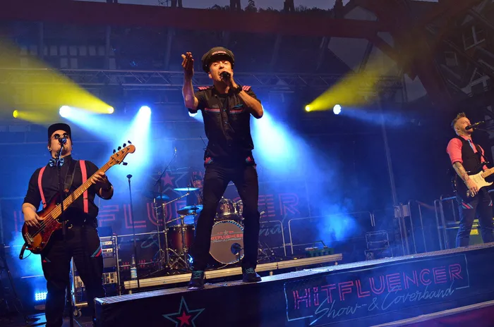 Die Band Hitfluencer tritt auf einer Bühne unter blauem und gelbem Scheinwerferlicht auf, mit dem Sänger in der Mitte und Musikern an Bass und Gitarre.