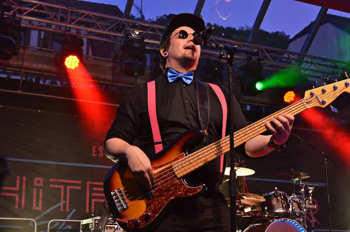 Ein Musiker der Band Hitfluencer mit Sonnenbrille, Kappe, blauer Fliege und rosa Hosenträgern spielt Bass und singt auf einer Bühne.