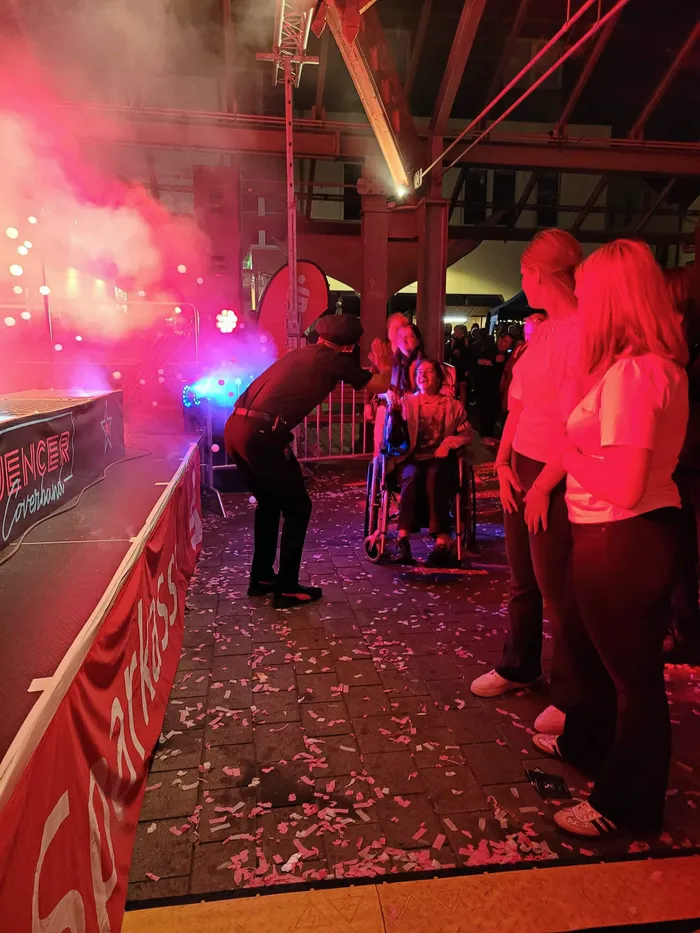 Ein Musiker beugt sich zu einer Person im Rollstuhl vor, während im Hintergrund eine Bühne mit rosa Licht und Rauch zu sehen ist.