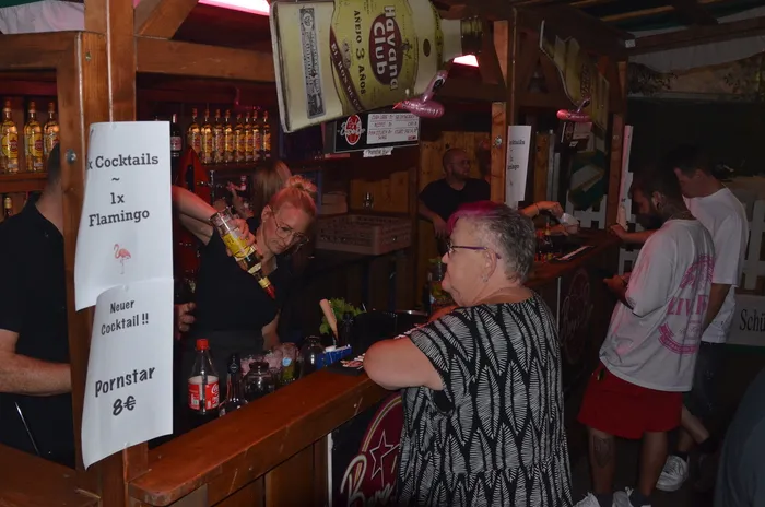 Eine Barkeeperin bereitet an einer Holzbar in einem Festzelt Cocktails für Gäste zu, darunter eine Kundin mit kurzen rosa Haaren.