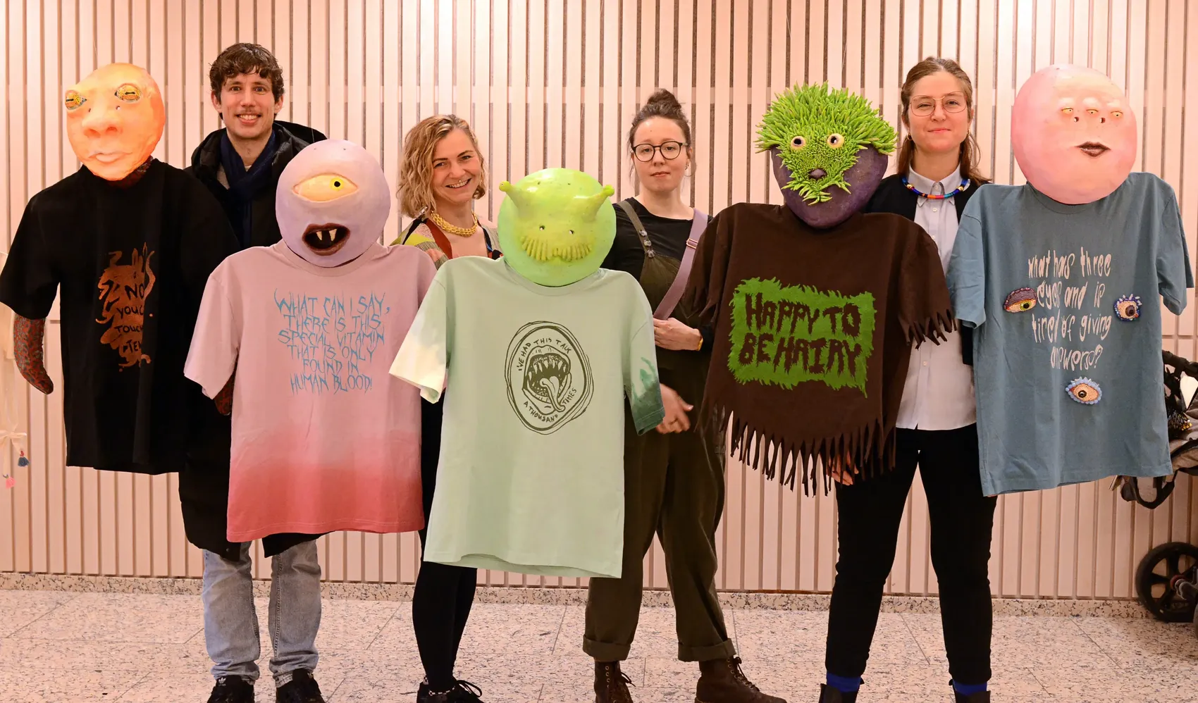 Vier Künstler stehen vor einer gerippten Wand und tragen übergroße, skurrile Masken, die Gesichter darstellen. Sie tragen Kleidung mit aufgedruckten Texten und Botschaften.