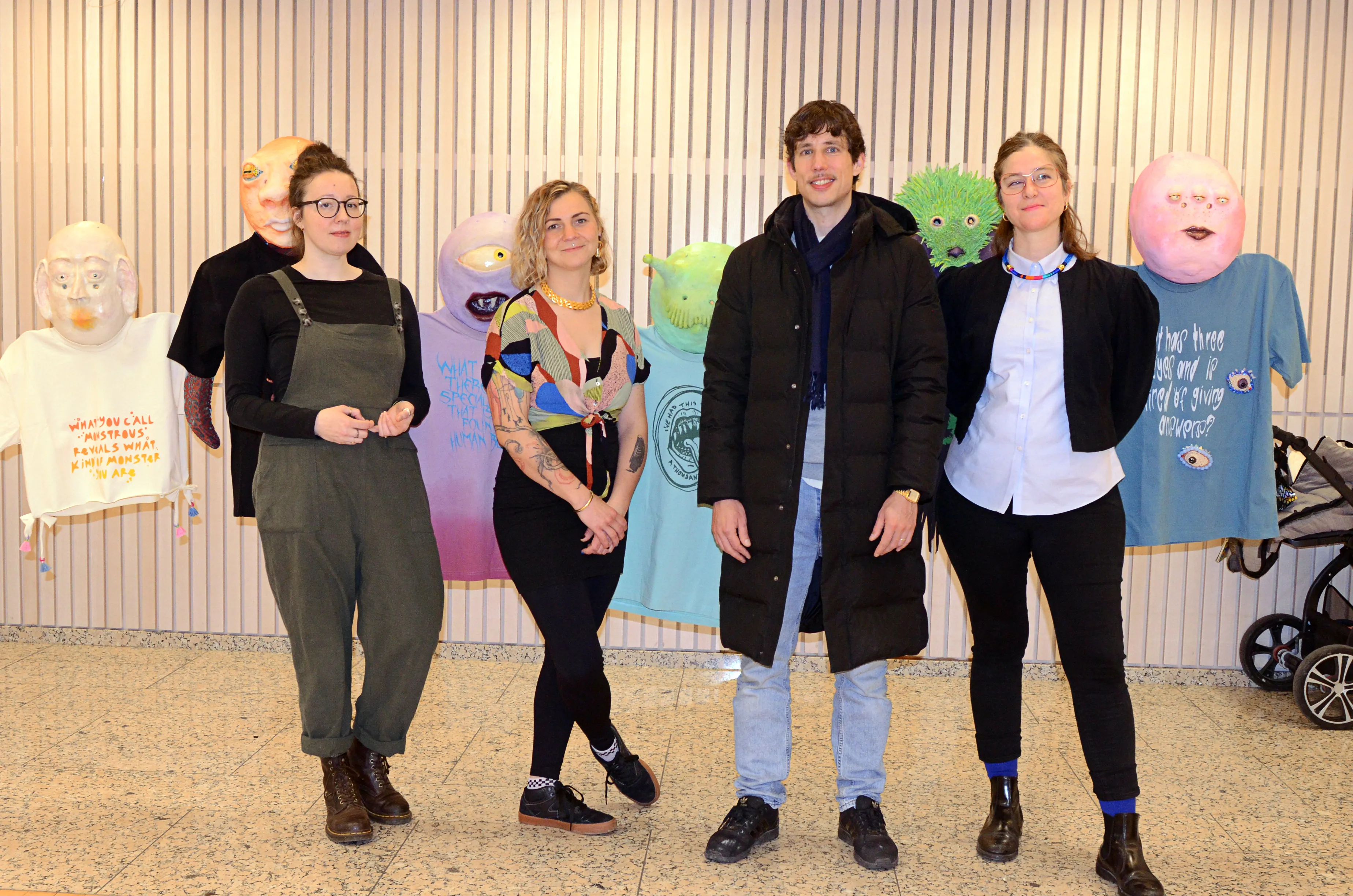 Vier junge Künstler stehen vor ihren farbenfrohen, skurrilen Figuren aus Stoff und anderen Materialien. Im Hintergrund eine Wand mit vertikalen Lamellen.