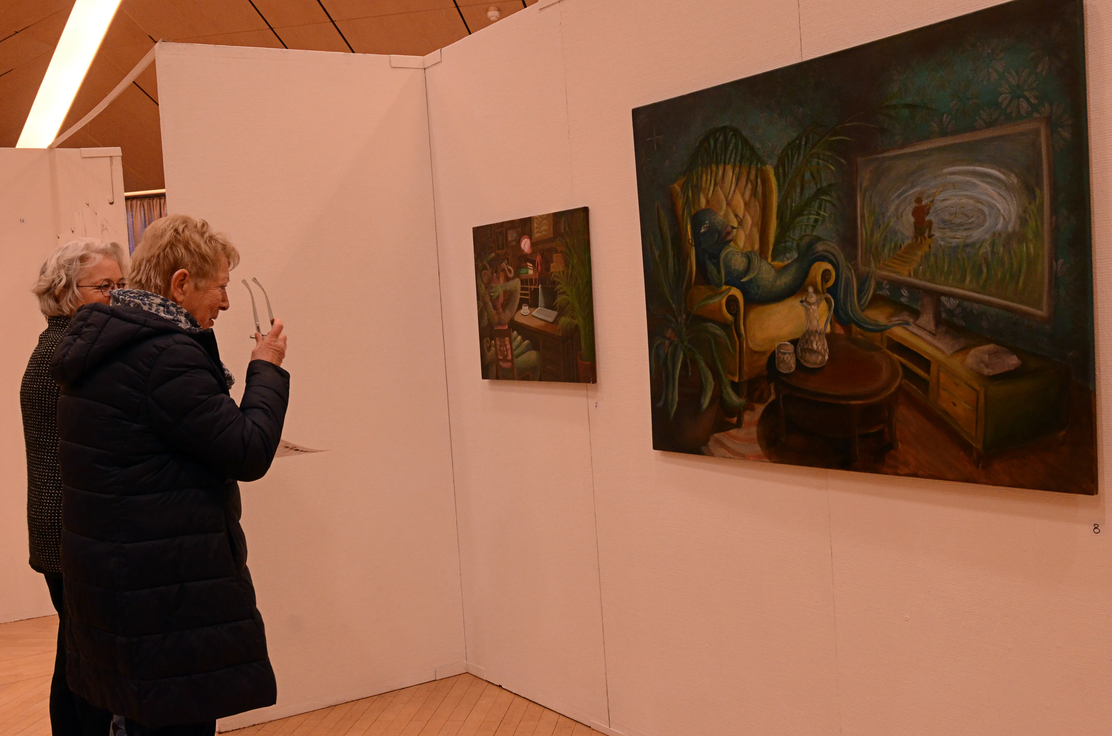 Zwei Frauen betrachten ein Gemälde in einer Ausstellung. Das Bild zeigt eine surreale Wohnszene mit einem blauen Wesen, das auf einem Sessel sitzt und einem Fernseher zusieht.