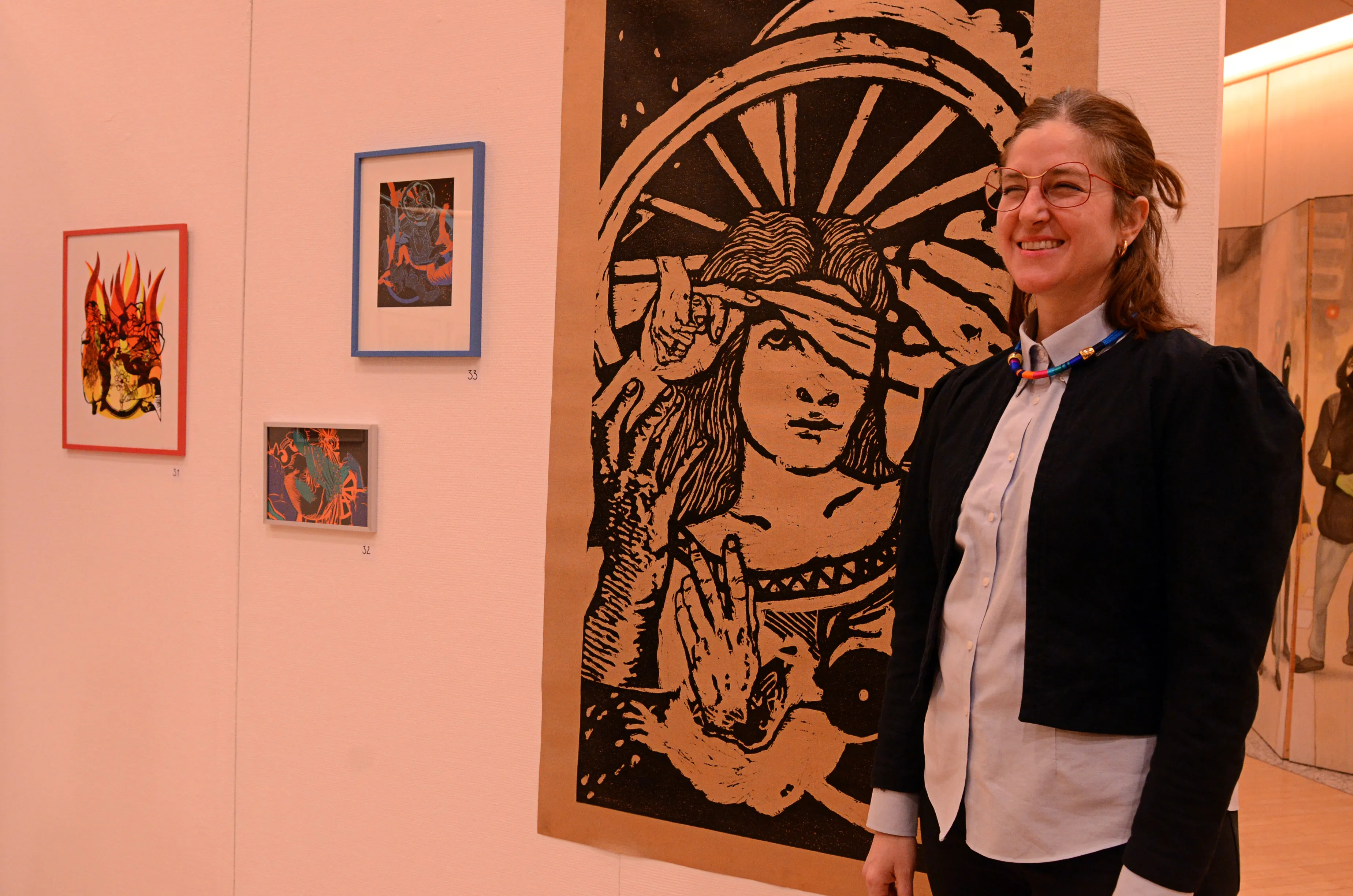 Eine Frau mit Brille und schwarzer Jacke lächelt neben einem großen, grafischen Druck mit dem Porträt einer Person mit erhobener Hand. Im Hintergrund sind weitere gerahmte Kunstwerke an einer hellen Wand zu sehen.