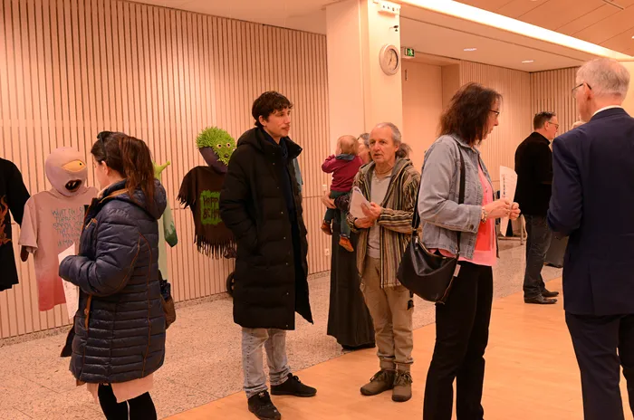 Eine Gruppe von Menschen steht in einem hellen Flur vor einer Tür und betrachtet Kunstwerke. Eine Person hält ein kleines Kind auf dem Arm, während eine andere eine Puppe trägt.