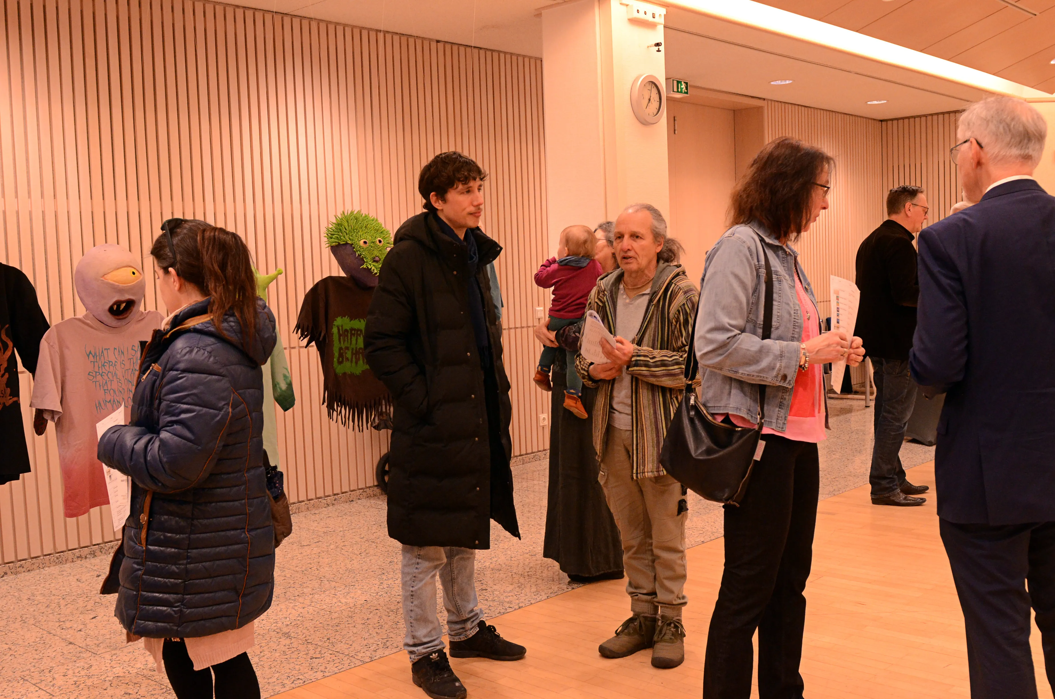 Eine Gruppe von Menschen steht in einem hellen Flur vor einer Tür und betrachtet Kunstwerke. Eine Person hält ein kleines Kind auf dem Arm, während eine andere eine Puppe trägt.