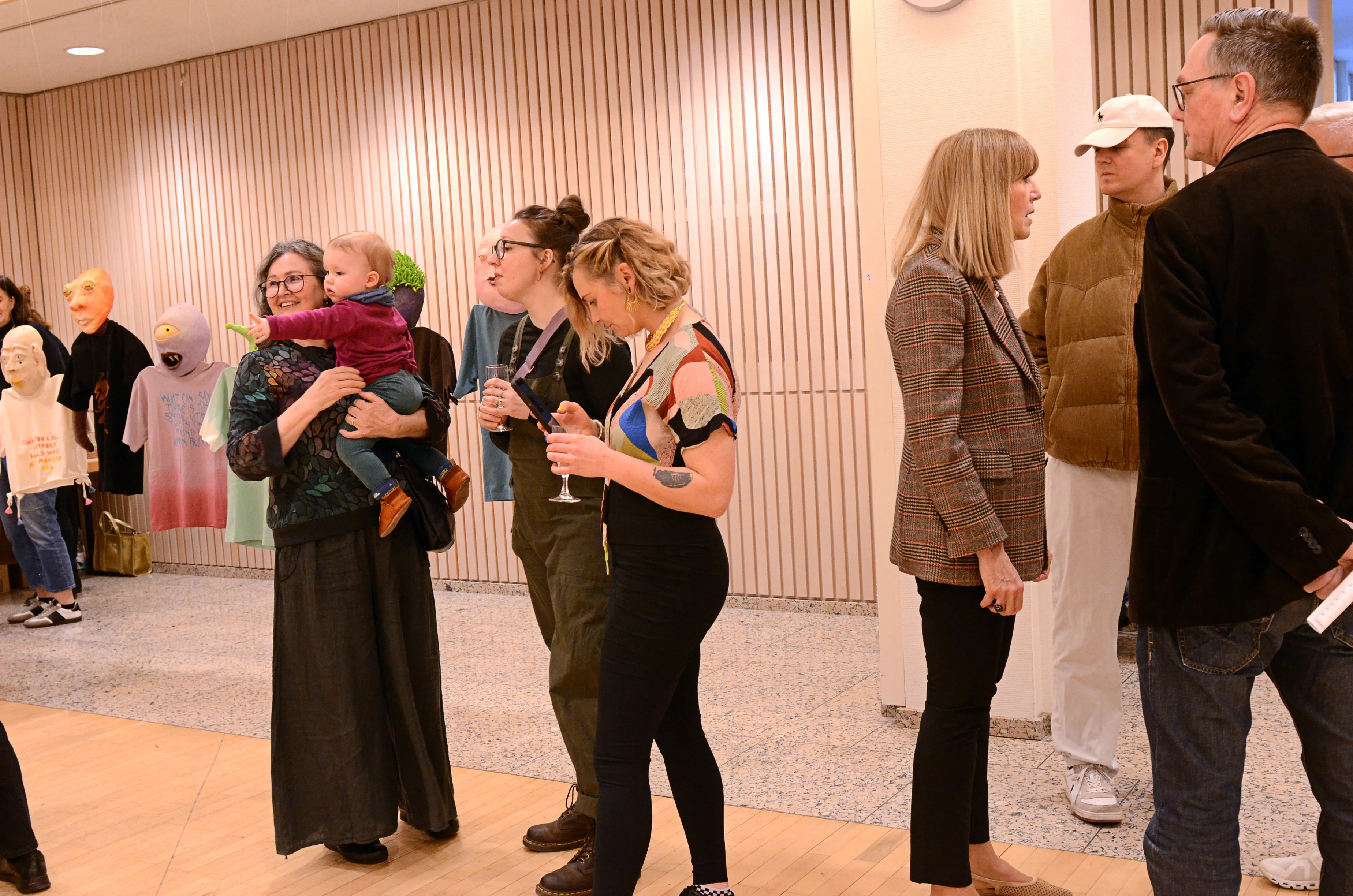 Eine Gruppe von Menschen steht in einem hellen Raum vor einer Reihe von Kunstwerken. Einige tragen Masken, eine Frau hält ein Kind im Arm.