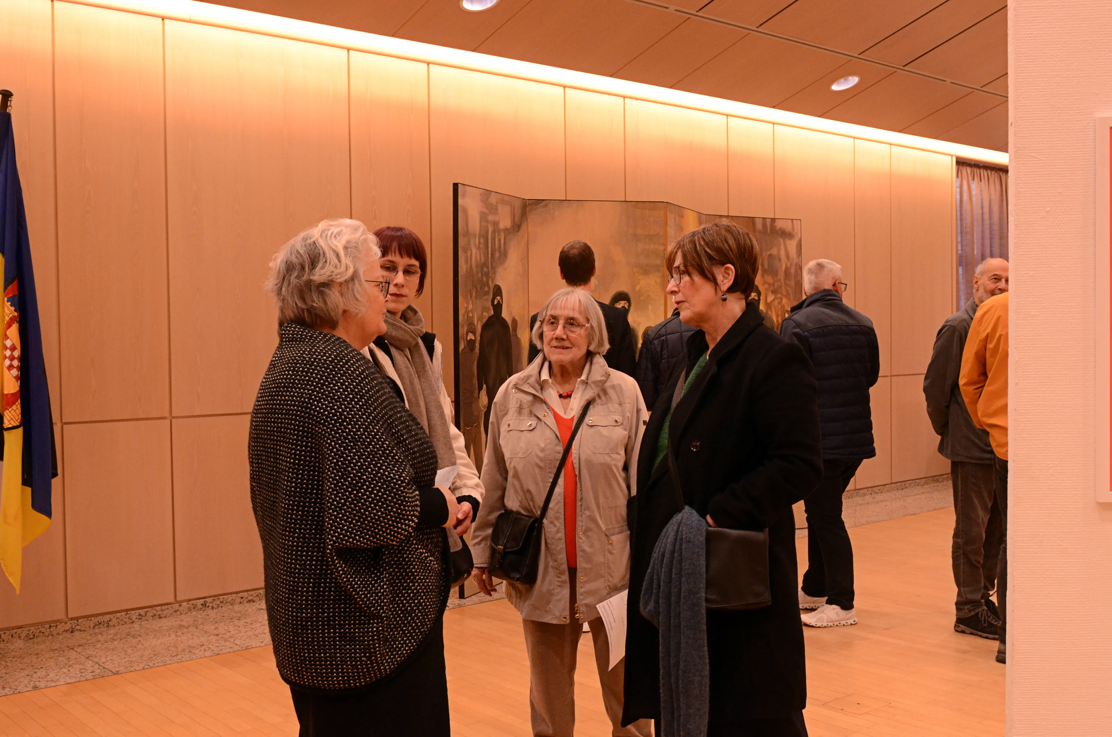 Mehrere Personen betrachten Kunstwerke in einer hellen Ausstellungshalle. Im Vordergrund stehen drei Frauen, die sich unterhalten, während im Hintergrund weitere Besucher und ein großes, gerahmtes Bild zu sehen sind.