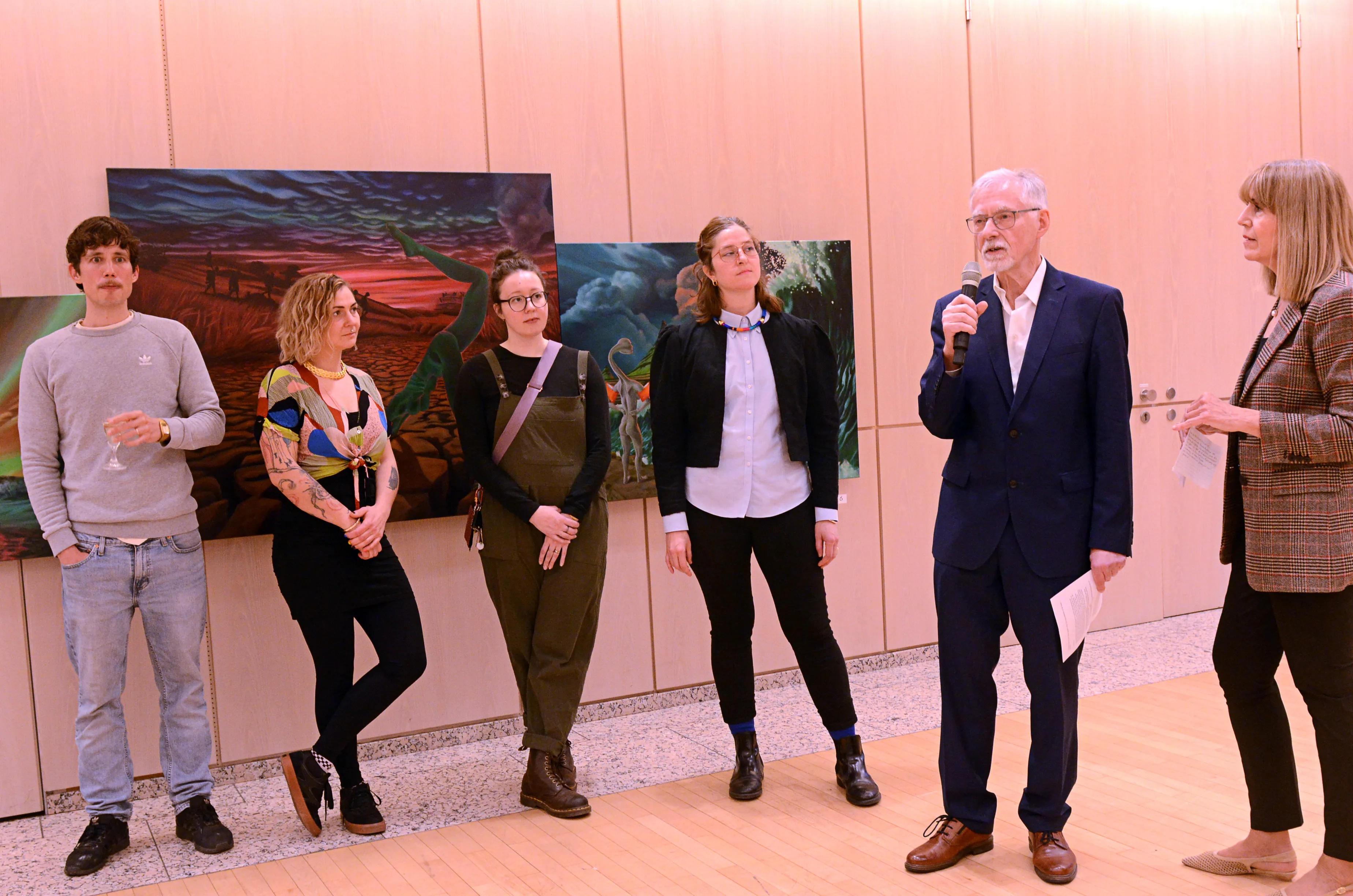 Fünf Personen stehen vor Gemälden in einem hellen Raum. Ein Mann mit Mikrofon spricht, während die anderen zusehen.