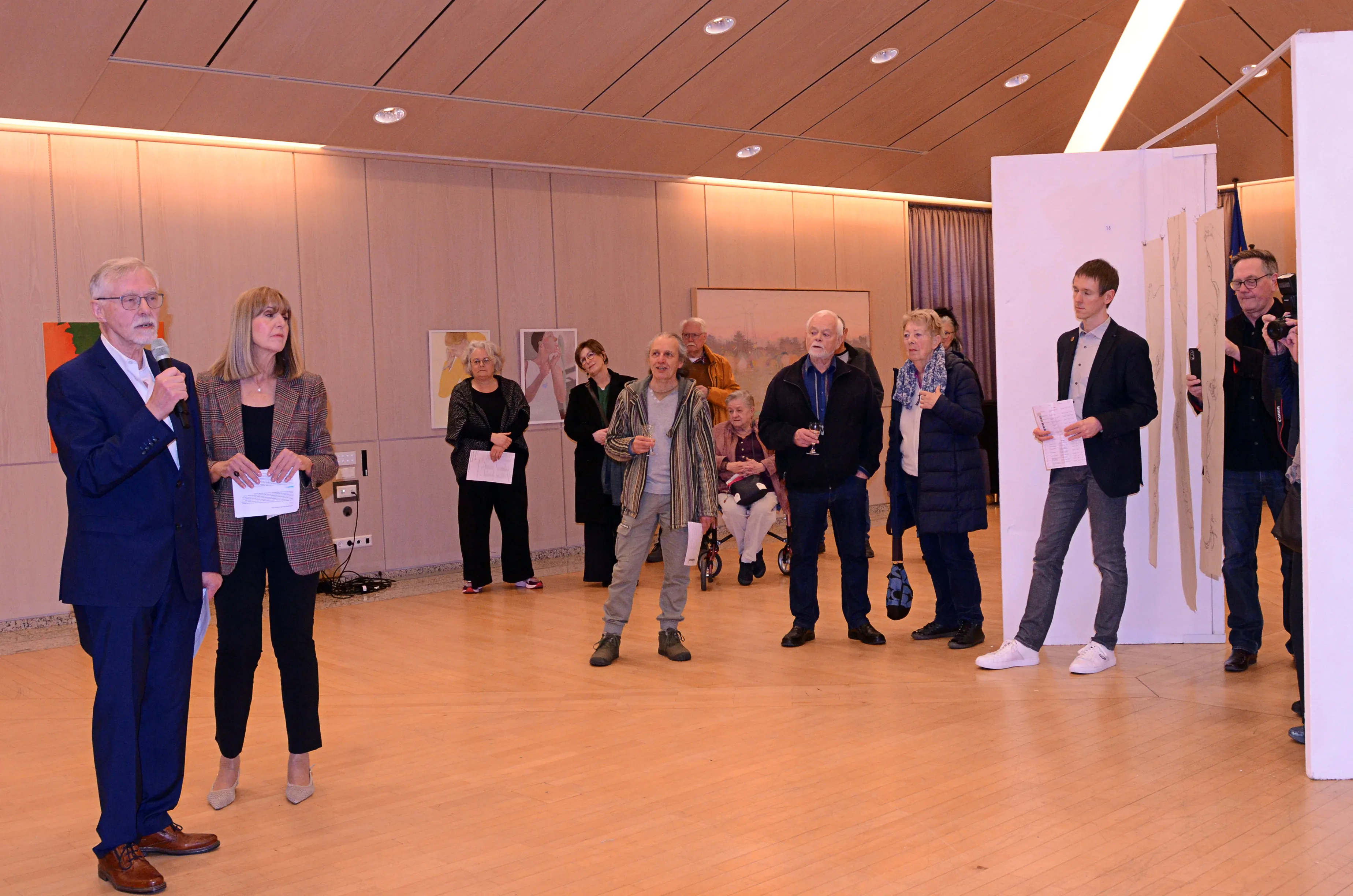 Eine Frau und ein Mann sprechen vor einer Gruppe von Menschen in einem hellen Raum. Im Hintergrund hängen Kunstwerke an den Wänden.