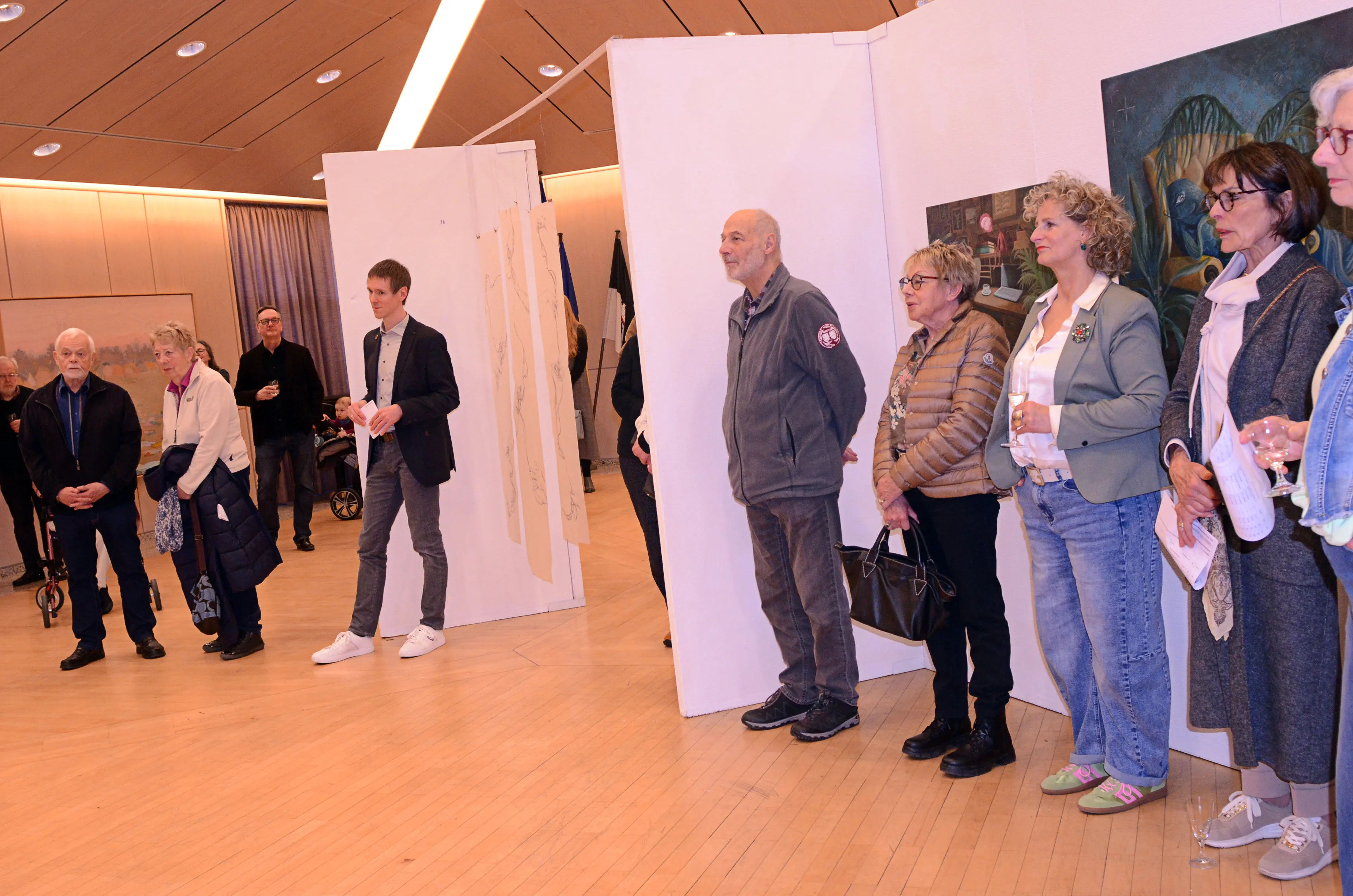 Besucher stehen vor Kunstwerken in einem hellen Raum, einige halten Gläser und betrachten die Ausstellung im Ratssaal.