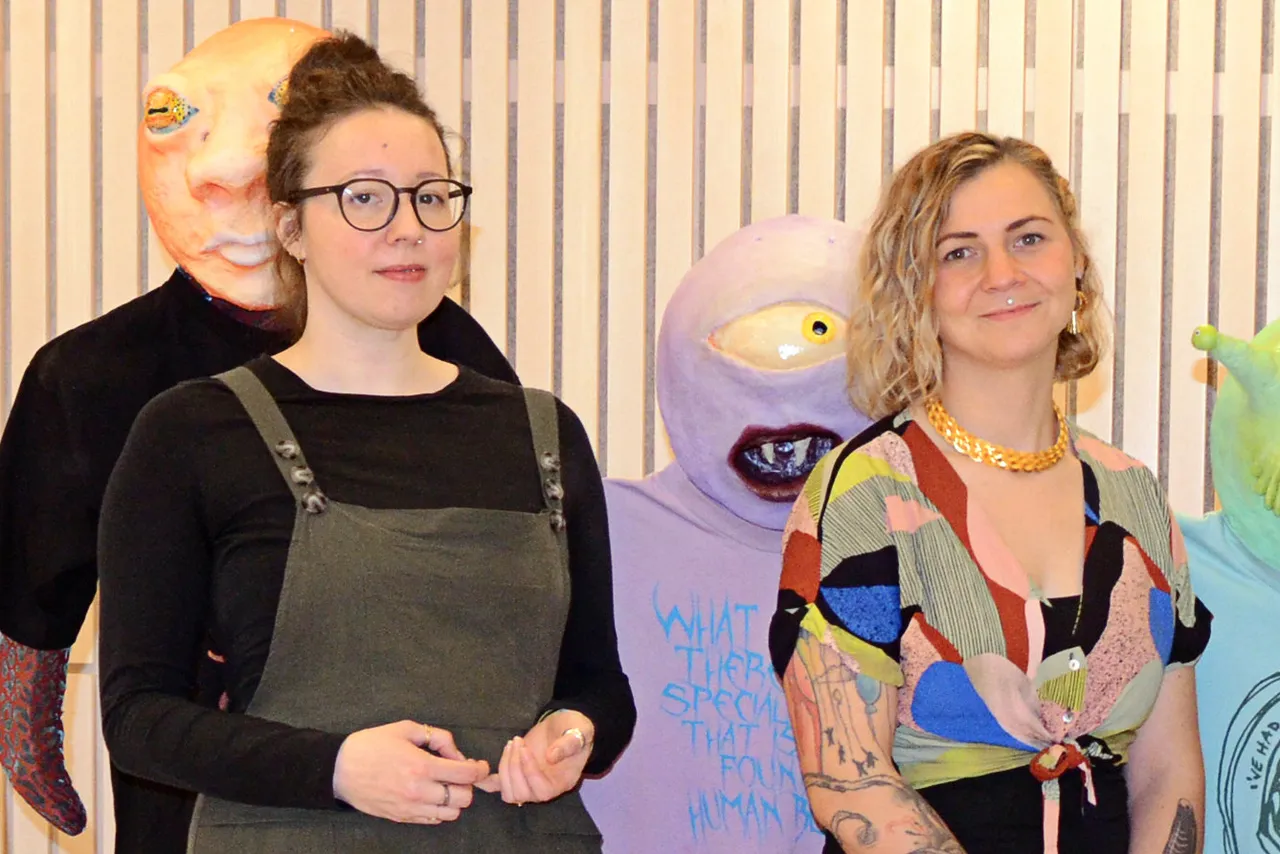 Zwei Frauen stehen vor großen, farbenfrohen Masken. Links Helena Zubler mit dunkler Kleidung und Brille, rechts Anja Herzog mit gemustertem Oberteil und goldenem Schmuck.
