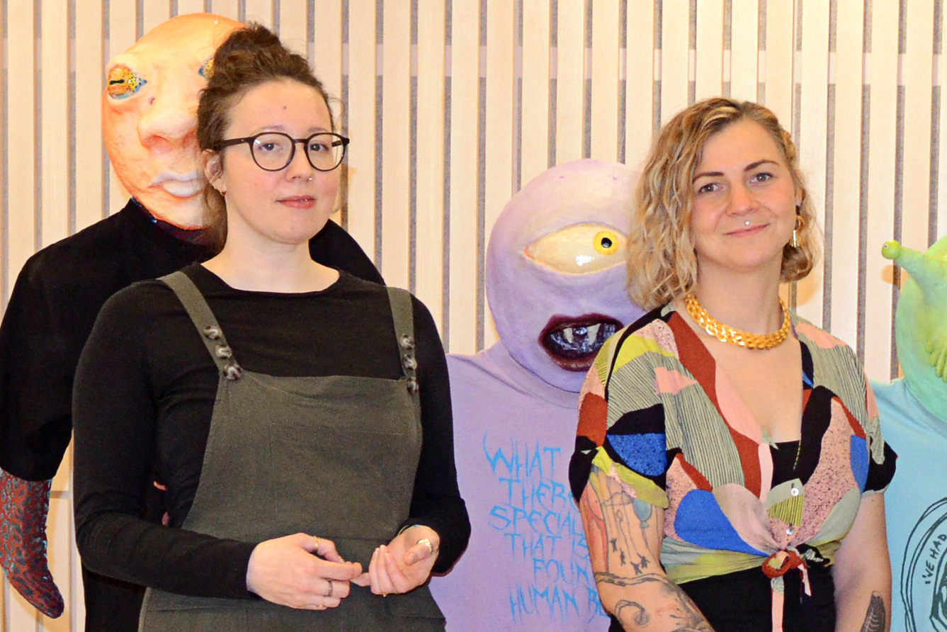 Zwei Frauen stehen vor großen, farbenfrohen Masken. Links Helena Zubler mit dunkler Kleidung und Brille, rechts Anja Herzog mit gemustertem Oberteil und goldenem Schmuck.