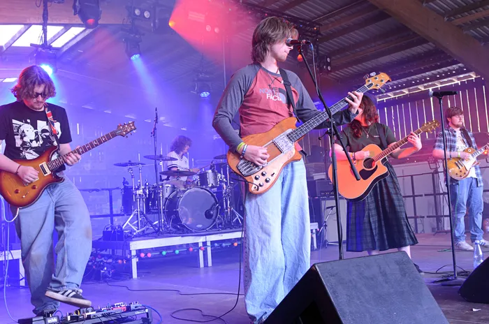 Eine Band spielt auf einer Bühne vor Publikum. Ein Mann mit langem Haar spielt Bass, während zwei weitere Musiker Gitarre spielen und ein Schlagzeuger im Hintergrund sitzt.
