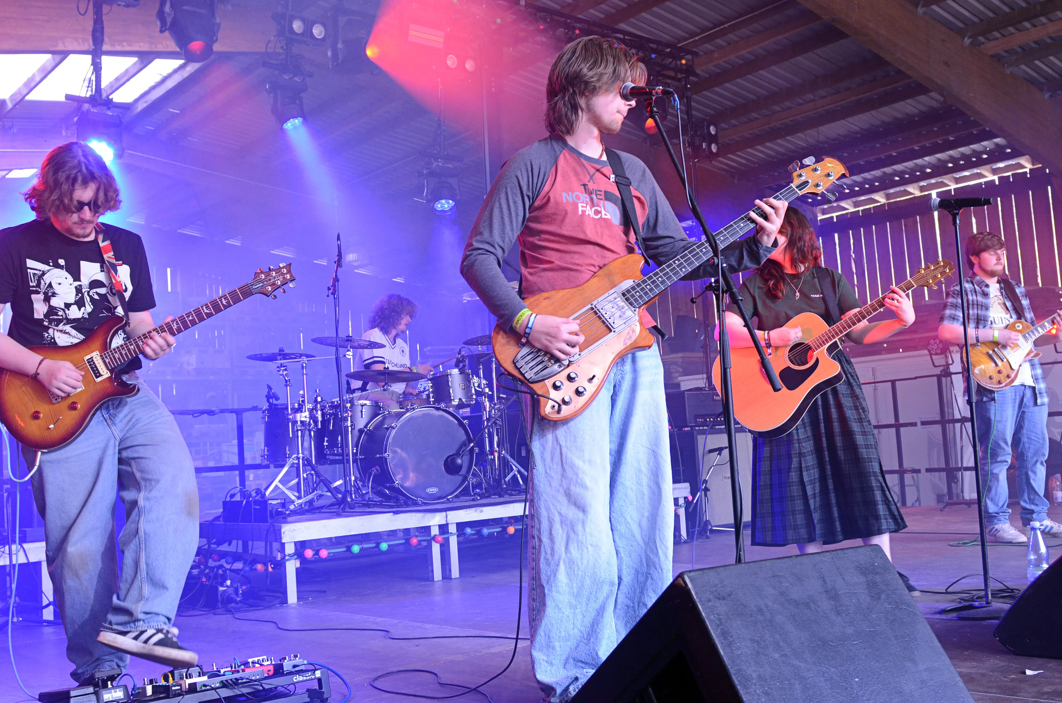 Eine Band spielt auf einer Bühne vor Publikum. Ein Mann mit langem Haar spielt Bass, während zwei weitere Musiker Gitarre spielen und ein Schlagzeuger im Hintergrund sitzt.
