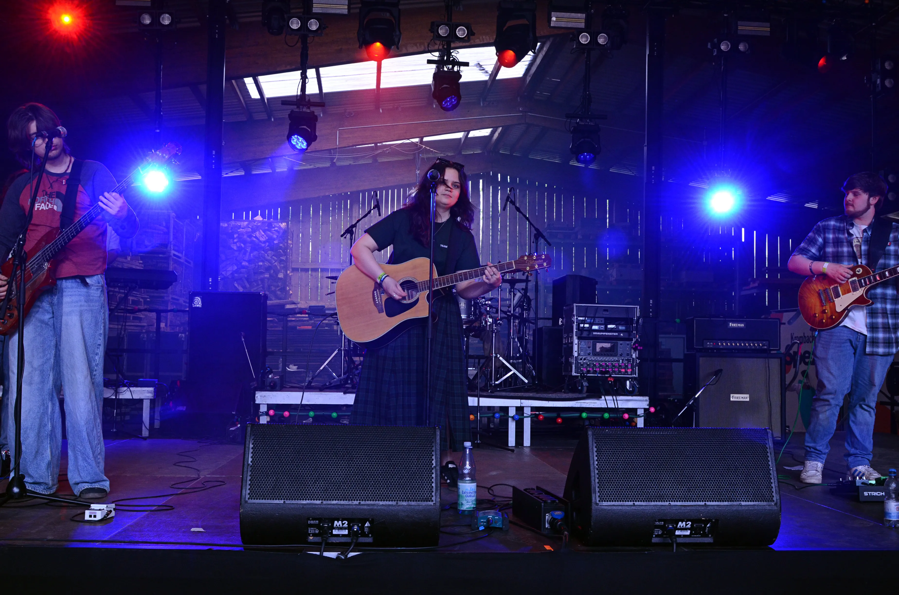 Eine Band spielt auf einer Bühne im Freien. Im Zentrum steht eine Person mit Gitarre, flankiert von Bassist und Gitarrist, während Bühnenlichter in Rot und Blau für Stimmung sorgen.
