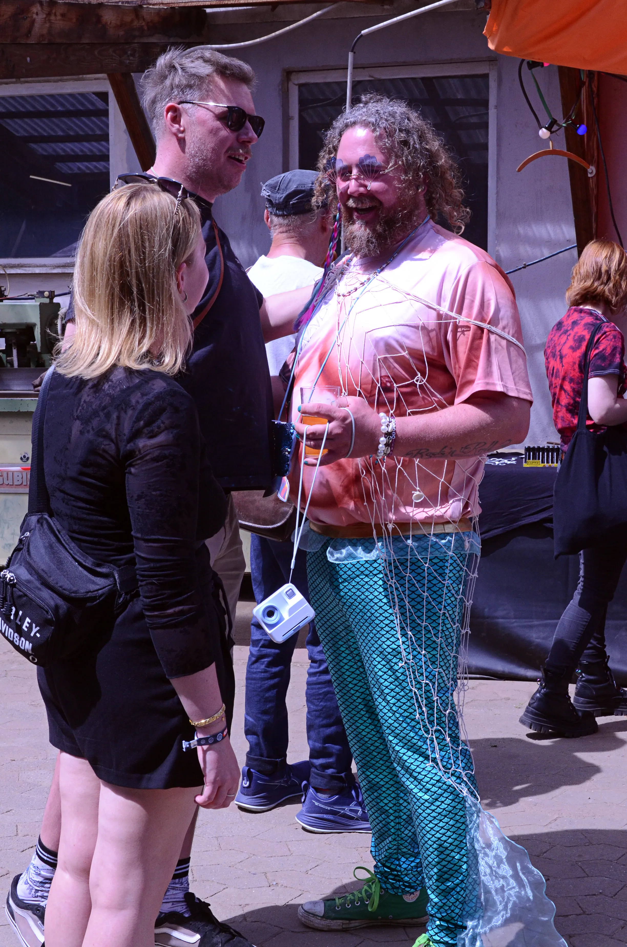 Zwei Personen unterhalten sich mit einem Mann, der ein Fischernetz und eine Kamera um den Körper trägt. Im Hintergrund sind weitere Festivalbesucher zu sehen.